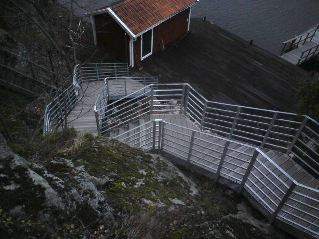 En lång, slingrande metalltrappa med kompositsteg, som leder nedför en bergsvägg till en brygga vid vattnet. Till vänster ses ett litet brygghus.