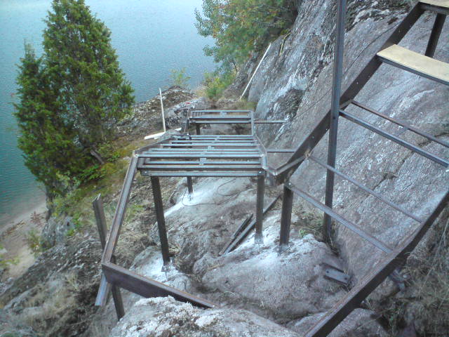 En lång, slingrande metalltrappa under konstruktion nedför en bergsvägg, som leder ner mot vattnet.