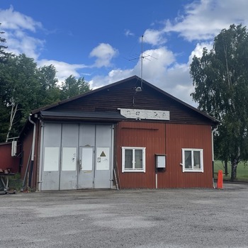 En verkstad sedd framifrån i landskapsvy. Fasaden är röd, med två fönster och en stor port i metall. Omgivningen består av en grusplan och till höger syns en byggbod.